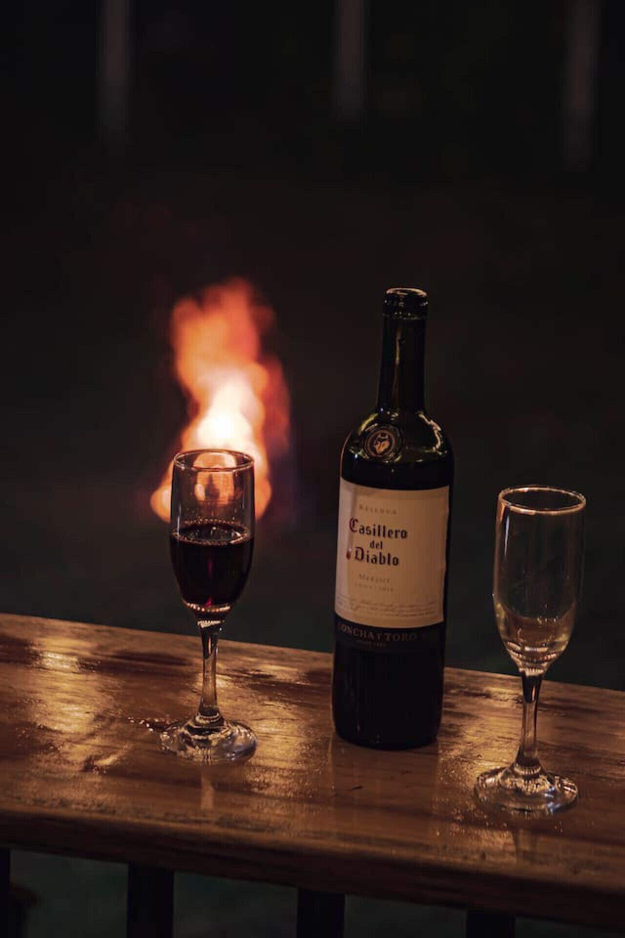 Una botella de vino Casillero del Diablo y dos copas sobre una superficie de madera, con un fuego al fondo.