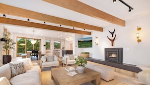 A spacious living room with a fireplace, wooden beams, and a large window offering a view of greenery. - Boulder Luxury: Panoramic Mountain Views, Cedar Hot Tub, Wellness and Romance! (Boulder)