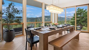 A modern dining area with a wooden table set for four, large windows offering a mountain view, and a potted plant on the left. - Boulder Luxury: Panoramic Mountain Views, Cedar Hot Tub, Wellness and Romance! (Boulder)