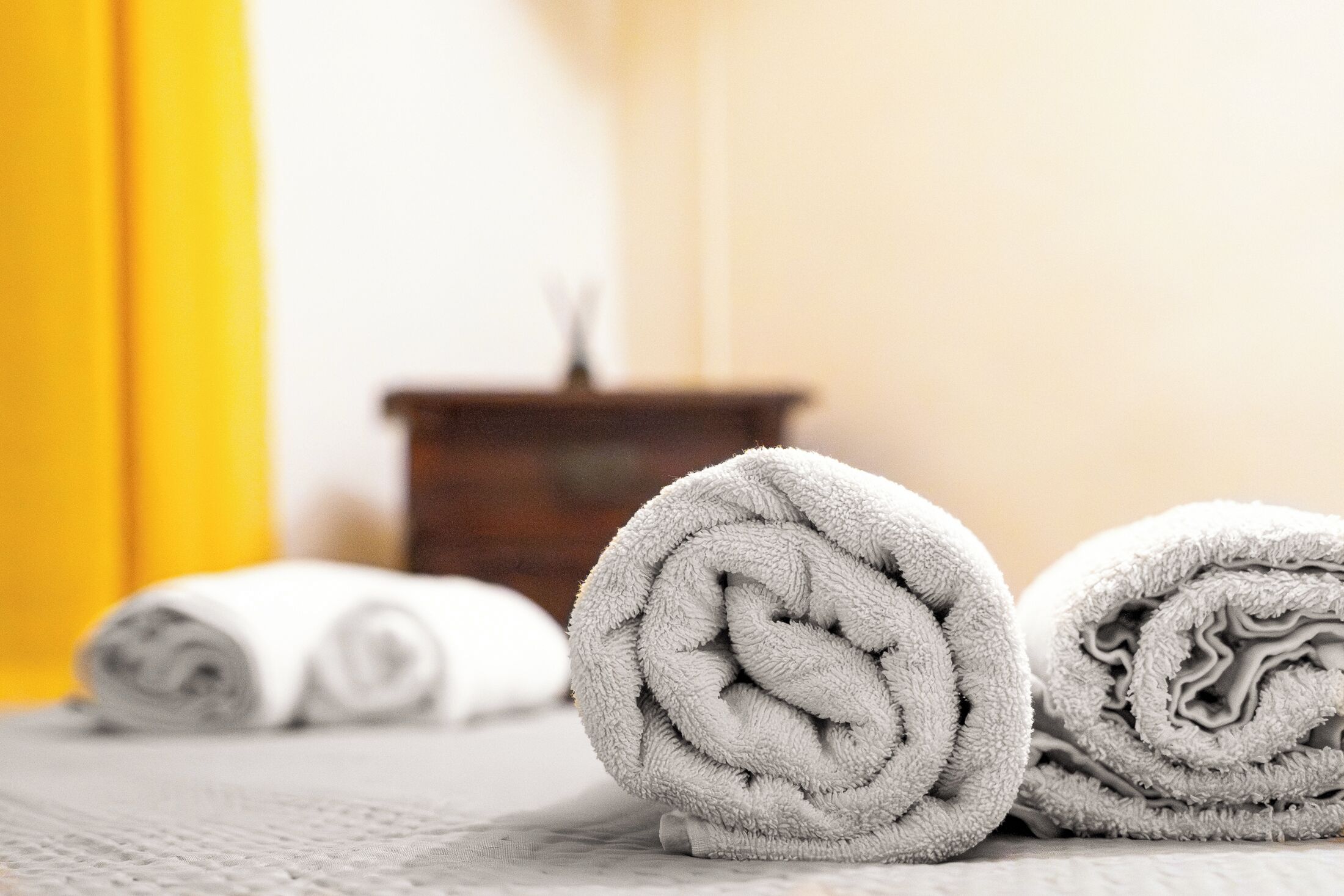 Three rolled towels on a textured surface, with a wooden side table and a yellow wall in the background.
