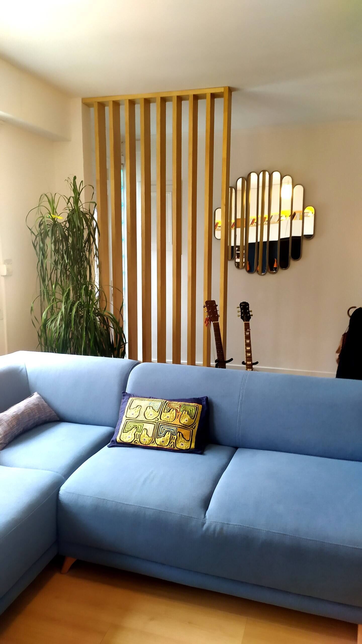 Ein blaues Sofa mit einem gemusterten Kissen, eine Trennwand aus Holzlatten, ein Topf mit Pflanze und zwei Gitarren an der Wand.