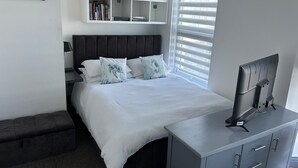 Un dormitorio moderno con cama, mesita de noche, televisor y ventana con persianas.