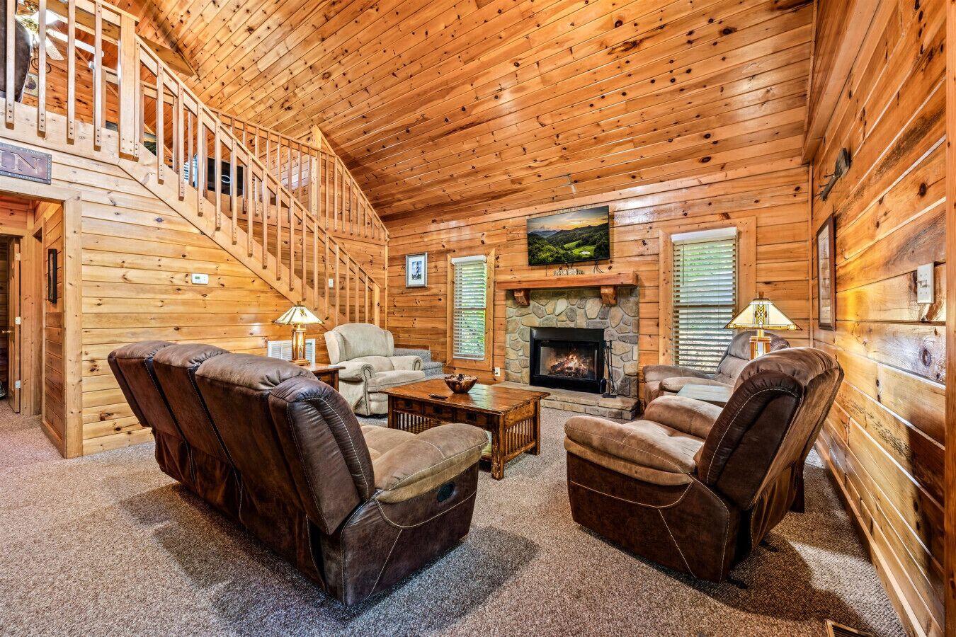 A log cabin interior with a fireplace, leather chairs, a sofa, and a staircase.