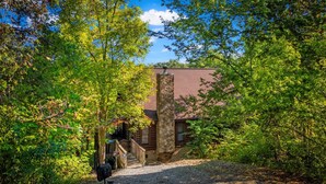 A rustic log cabin with a stone chimney, surrounded by dense foliage and a gravel path leading to the entrance.