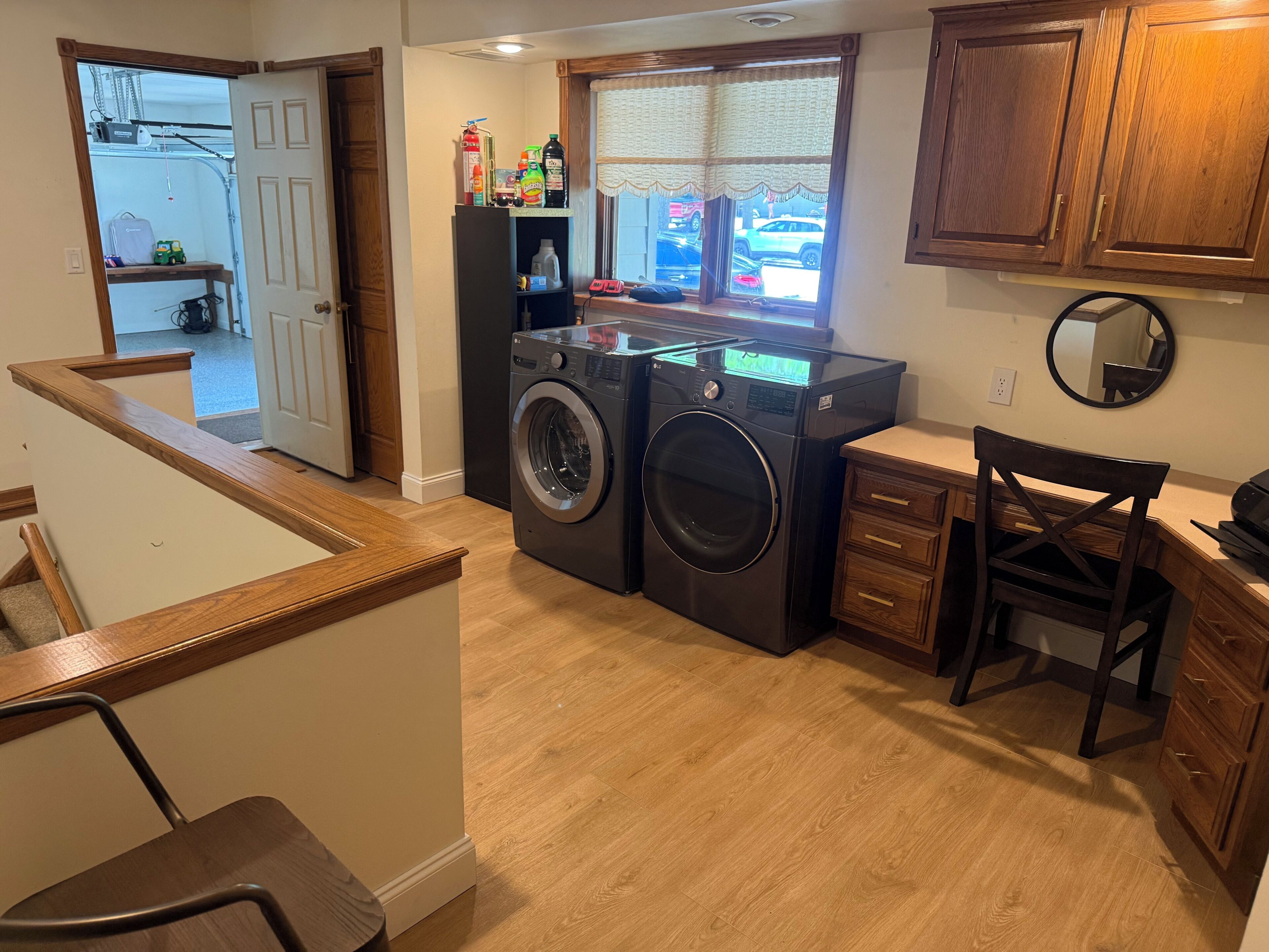 A laundry area with a washer and dryer, wooden cabinets, a chair, and a window with a view of parked cars.