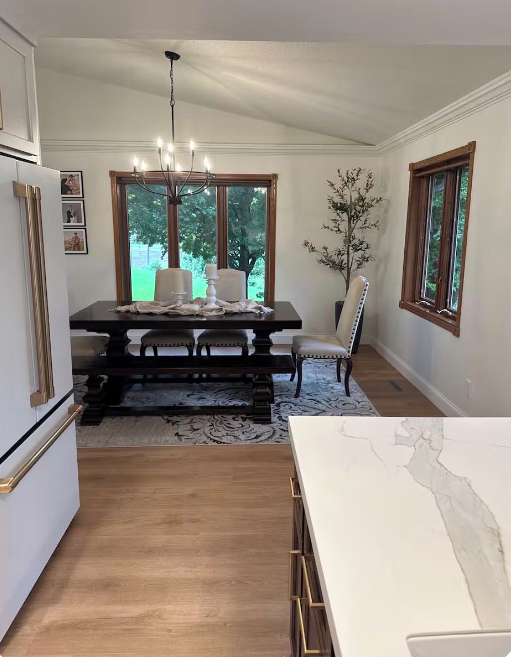 A dining area with a dark wooden table, white chairs, and a chandelier. There are windows with views of greenery outside.