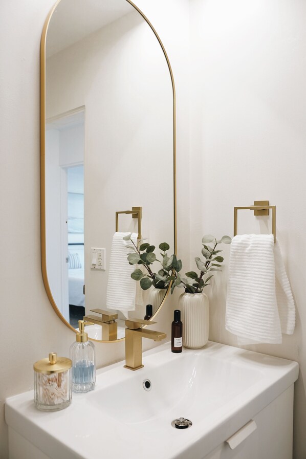 Un baño con lavamanos blanco, detalles dorados, un espejo grande y un perchero para toallas.