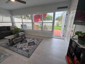 A living room with a sofa, coffee table, and a rug. There is a bookshelf with books and a plant. The room has large windows and a door leading outside.