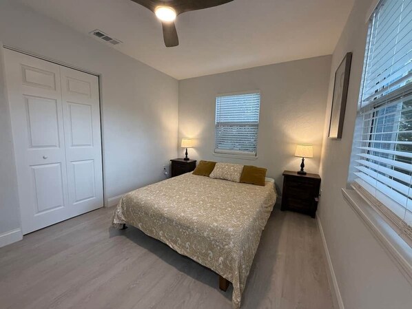 A bedroom with a bed, nightstands, a ceiling fan, a window with blinds, and a door.