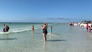 Escena de playa con gente en el agua y en la arena, cielos despejados y un mar tranquilo.