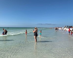 Una spiaggia con persone in acqua e sulla sabbia, cielo sereno e mare calmo.