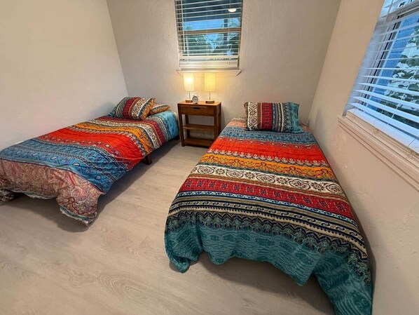 A small room with two single beds, a nightstand with a lamp, and a window with blinds.