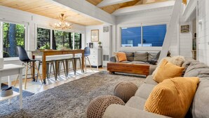 A spacious living room with a wooden ceiling, a dining area with a wooden table and chairs, and a large window offering a view of the outdoors. - Lake Winnipesaukee Cottage | Beach + Paddleboards (Meredith)