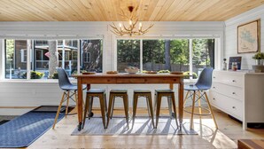 A dining area with a wooden table and four chairs, large windows, and a chandelier. - Lake Winnipesaukee Cottage | Beach + Paddleboards (Meredith)