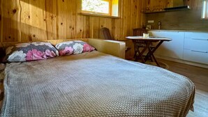 A wooden cabin bedroom with a bed, pillows, a window, a small table, and a kitchenette.