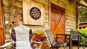A rustic log cabin with a dartboard on the wall, a wooden door, and a stone patio with a folding chair and table.