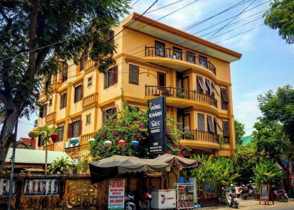 A multi-story yellow building with balconies, a sign for 'Dong Khánh House', and a small outdoor seating area with a canopy.
