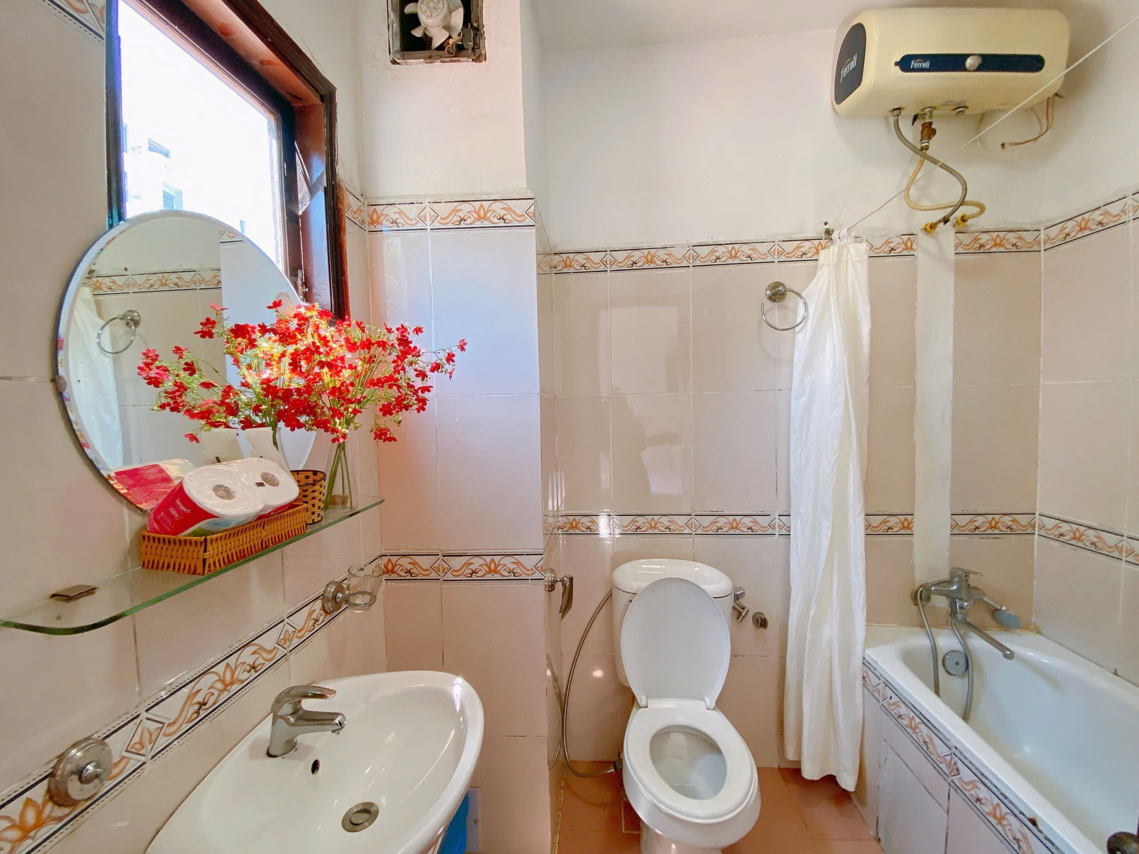 Ein Badezimmer mit Waschbecken, Toilette und Badewanne. Ein Spiegel mit dekorativem Rahmen, ein Ablagefach mit Pflegeprodukten und eine Blumenkomposition sind ebenfalls vorhanden.