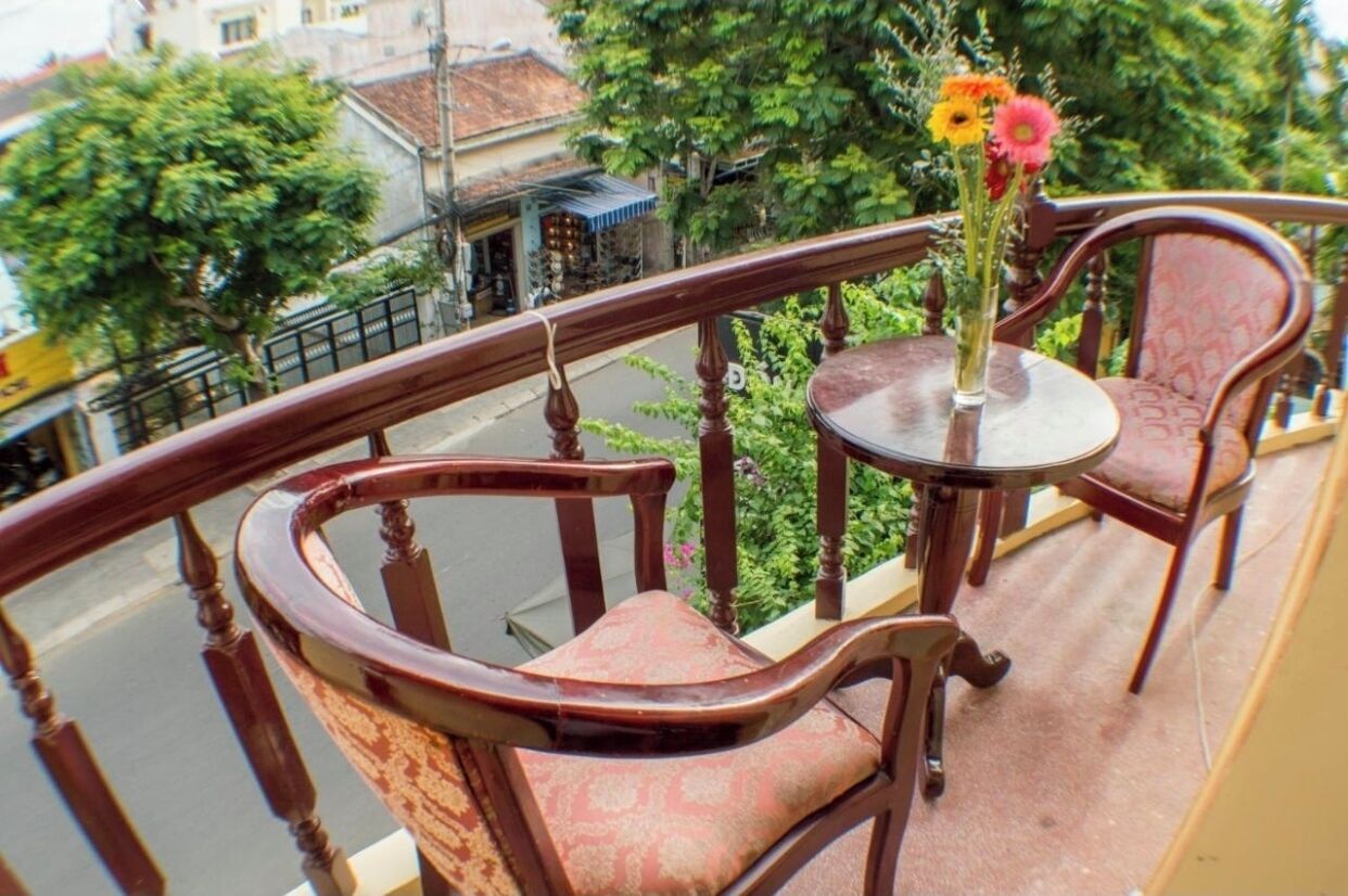A balcony with wooden chairs and a table, a vase with flowers, and a view of the street and buildings.