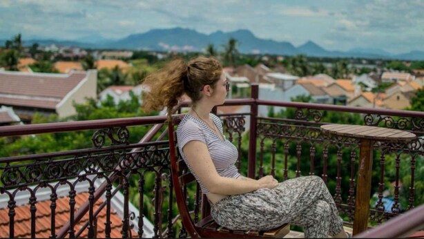 Eine Frau sitzt auf einem Balkon mit Blick auf eine Wohngegend und Berge im Hintergrund.