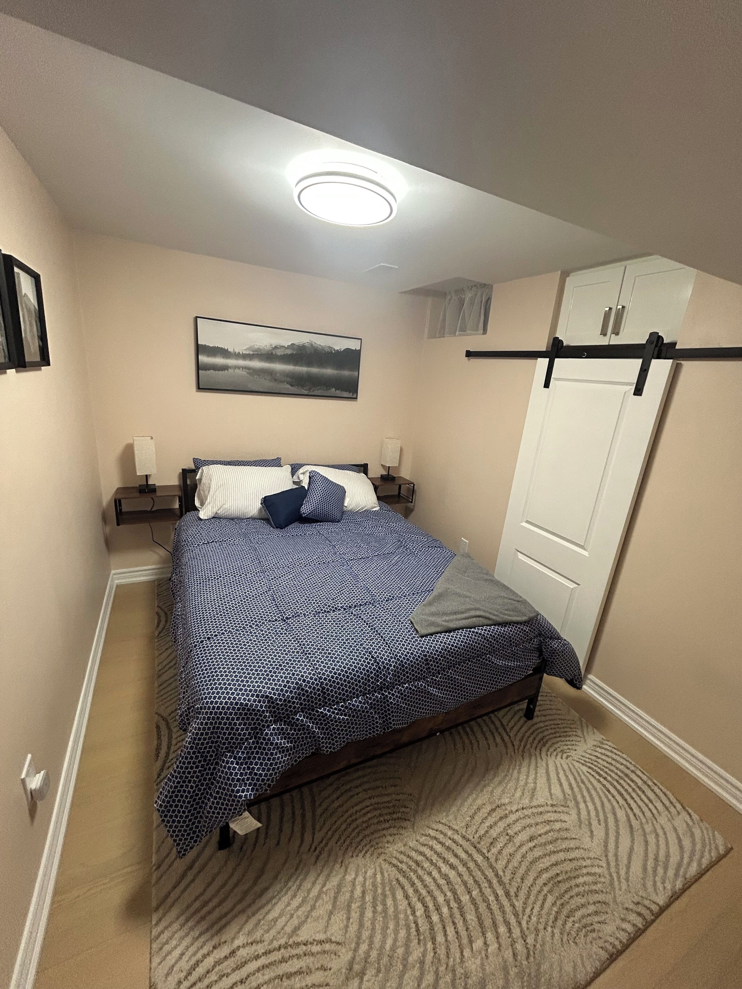 Ein Schlafzimmer mit Bett, Nachttisch, Lampe, einem Wandbild, einer Tür und einer Deckenleuchte.