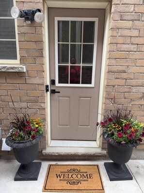 A door with a welcome mat, two flower pots, and a window with a light fixture. - 2-bedroom, 1-bathroom basement apartment, perfect for small families or couples. (Brampton)