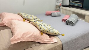 Un lit bien fait, avec une literie rose et beige, un coussin à motifs et une serviette enroulée sur un plancher en bois.
