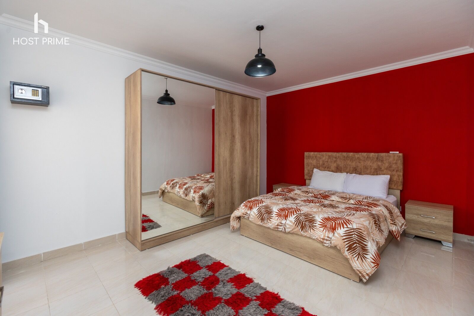 Un dormitorio con cama, mesita de noche, espejo, pared roja y luz de techo.