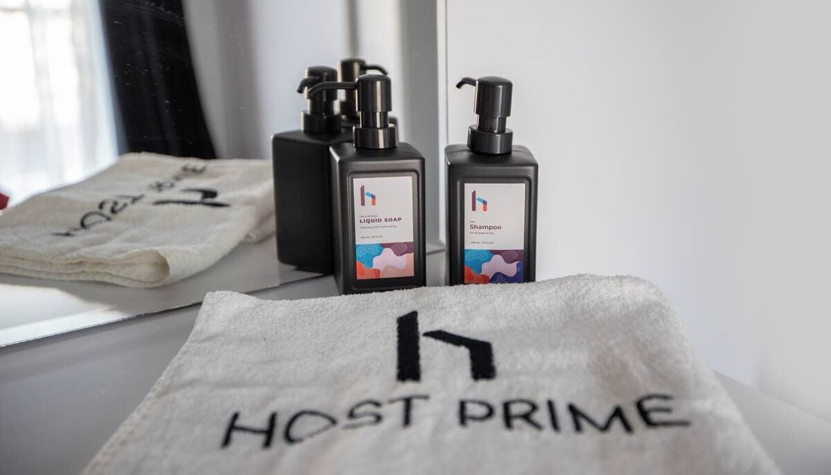 Casa de banho com toalha rotulada "HOST PRIME" e duas embalagens de gel de banho e champô.