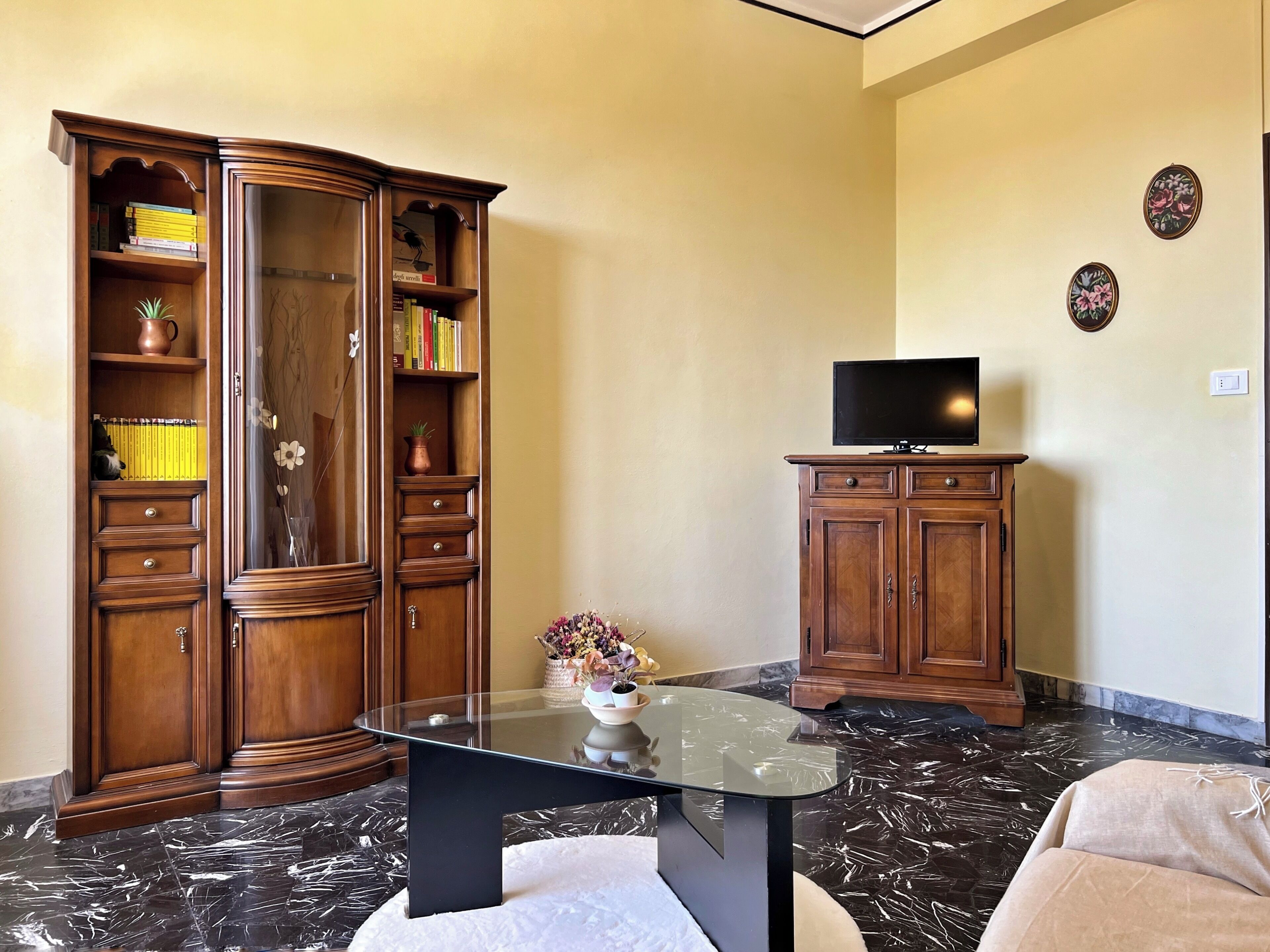 A living room with a glass-top coffee table, a wooden cabinet with books, a television on a stand, and a decorative vase.