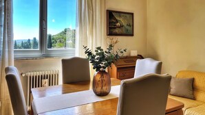 Een kamer met een houten tafel, stoelen, een vaas met planten, een schilderij aan de muur en uitzicht op groen buiten.