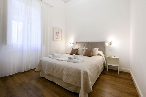 A neatly made bed with a beige bedspread and pillows, a bedside table with a lamp, and a framed picture on the wall.