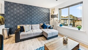 Ein modernes Wohnzimmer mit einem grauen Ecksofa, einem Holztisch und einem großen Fenster mit Blick auf Gebäude.