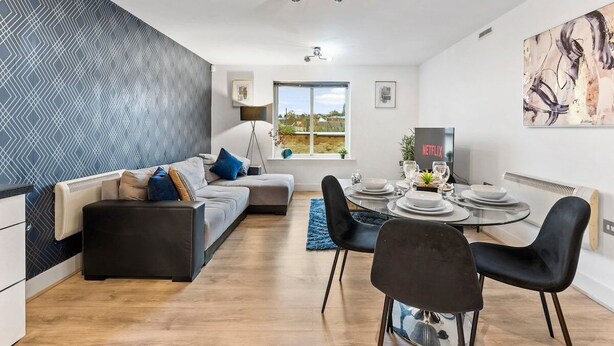 Ein modernes Wohnzimmer mit einer Couch, einem Esstisch mit Stühlen, einem großen Fenster und einem an der Wand befestigten Fernseher, der 'NETFLIX' zeigt.
