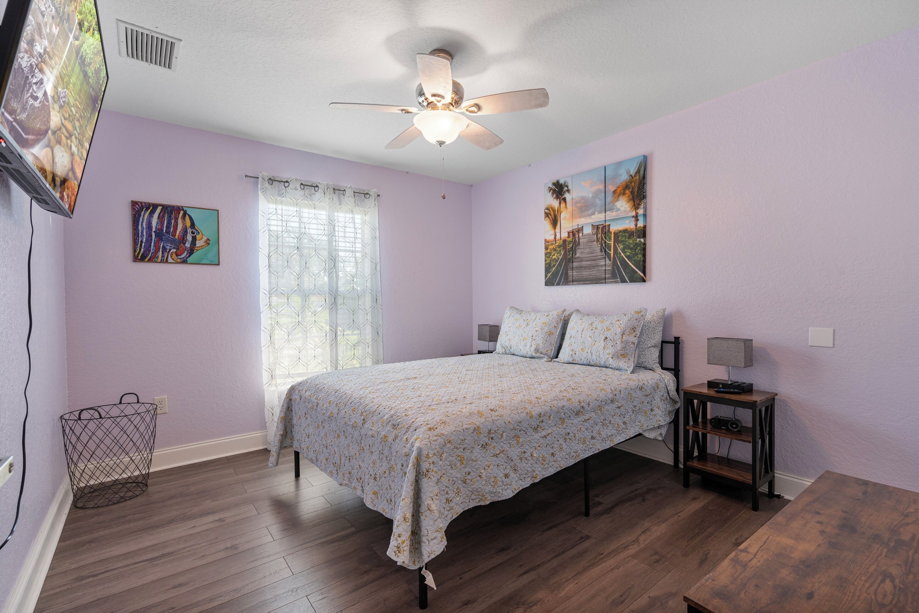 A bedroom with a bed, a ceiling fan, a nightstand, a lamp, a window with curtains, and a painting on the wall.