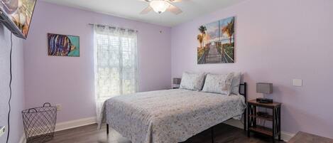 A bedroom with a bed, a ceiling fan, a nightstand, a lamp, a window with curtains, and a painting on the wall.