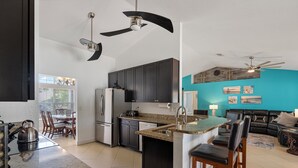 A modern kitchen with dark cabinets, a granite countertop, and a central island. There is a dining area with a table and chairs, and a living room with a black leather couch and a teal accent wall. - Pool Home in SW Florida. 4 bedrooms,2 bathrooms, close to shopping and beaches. (North Port)
