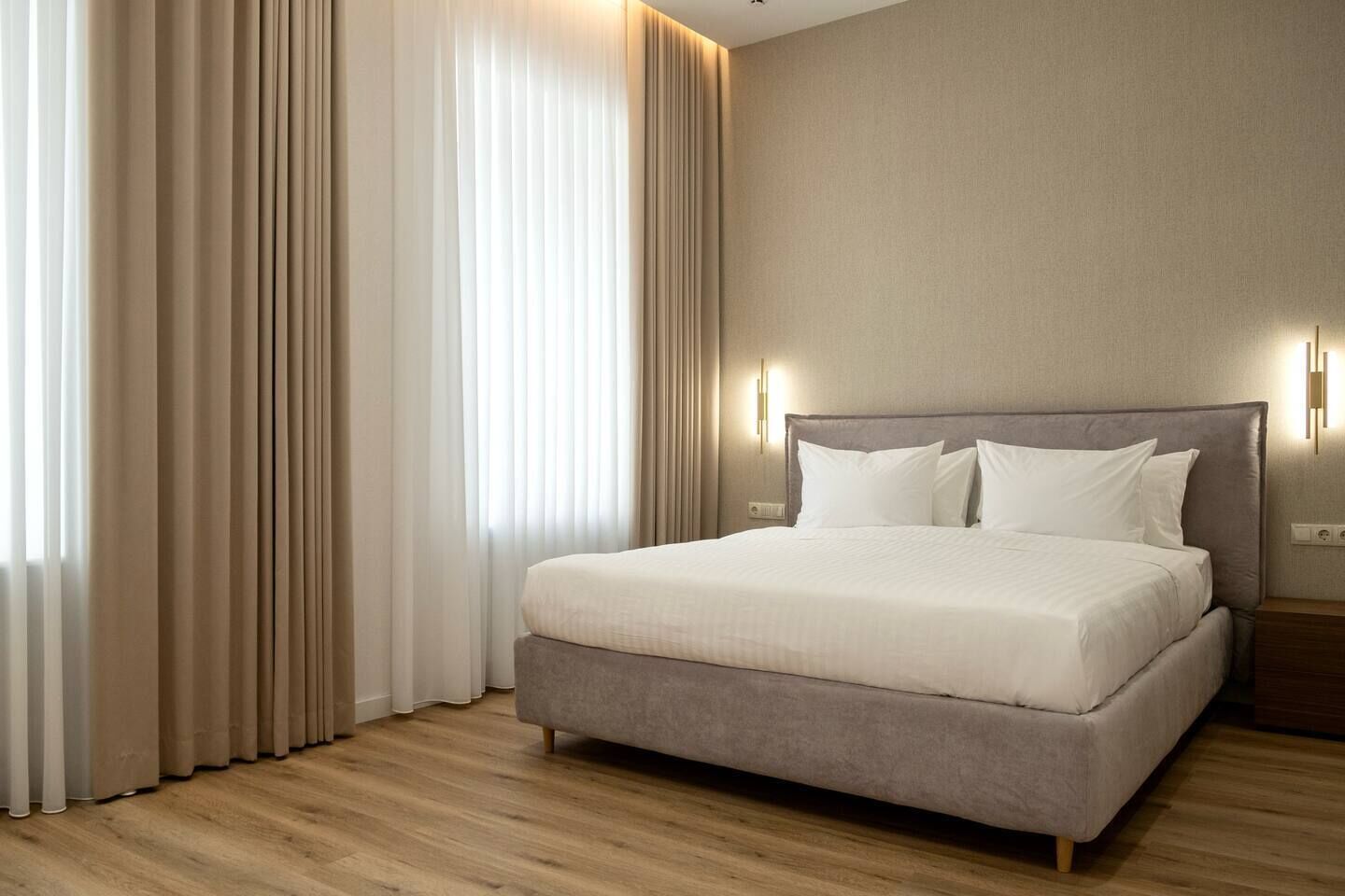 A modern hotel room with a large bed, light-colored curtains, and a wooden floor.