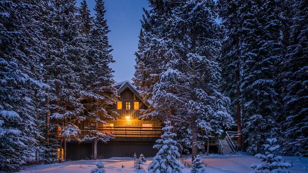 Una cabaña nevada de madera con entrada iluminada, rodeada de árboles cubiertos de nieve.