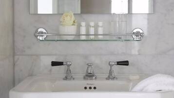 A bathroom with a white sink, two faucets, a glass shelf above the sink, and a mirror reflecting a white flower and bottles.