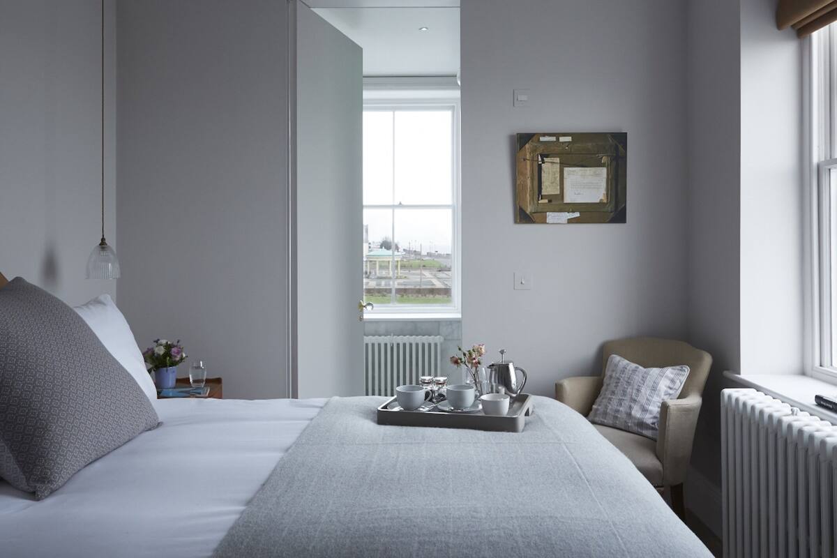 A modern hotel room with a large bed, a sitting area with a chair, a tray with breakfast items, and a window with a city view.