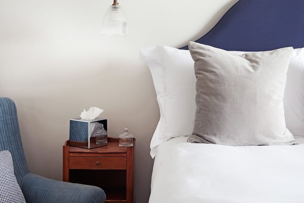A neatly made bed with white and gray pillows, a wooden nightstand with a tissue box and glass containers, and a blue armchair.