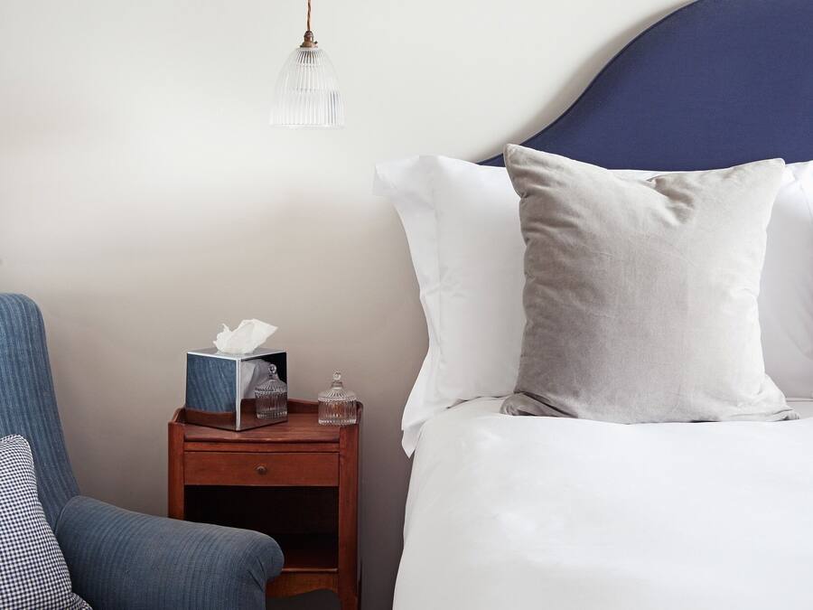 A neatly made bed with white and gray pillows, a wooden nightstand with a tissue box and glass containers, and a blue armchair.