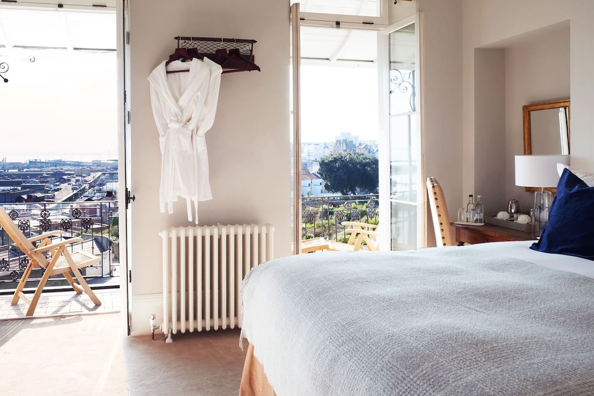 A bedroom with a large bed, a radiator, a wooden chair, and a view of the city through a large window.