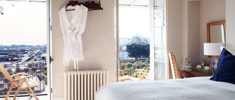 Une chambre avec un grand lit, un radiateur, une chaise en bois et une vue sur la ville par une grande fenêtre.