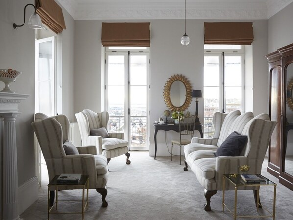 Amplia sala de estar con techo alto, ventanas grandes con persianas marrones y vista a la ciudad. Hay dos sillones, un sofá y una mesita de noche con una lámpara.