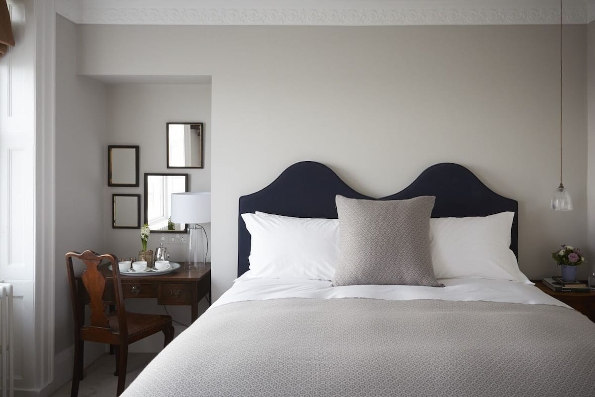 A neatly arranged bedroom with a large bed, a bedside table with a lamp, a chair, and a desk with a mirror and a vase of flowers.