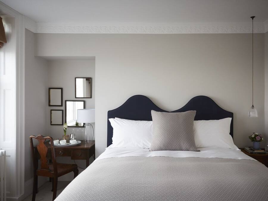 Un dormitorio ordenado con una cama grande, una mesita de noche con lámpara, una silla y un escritorio con espejo y un florero.
