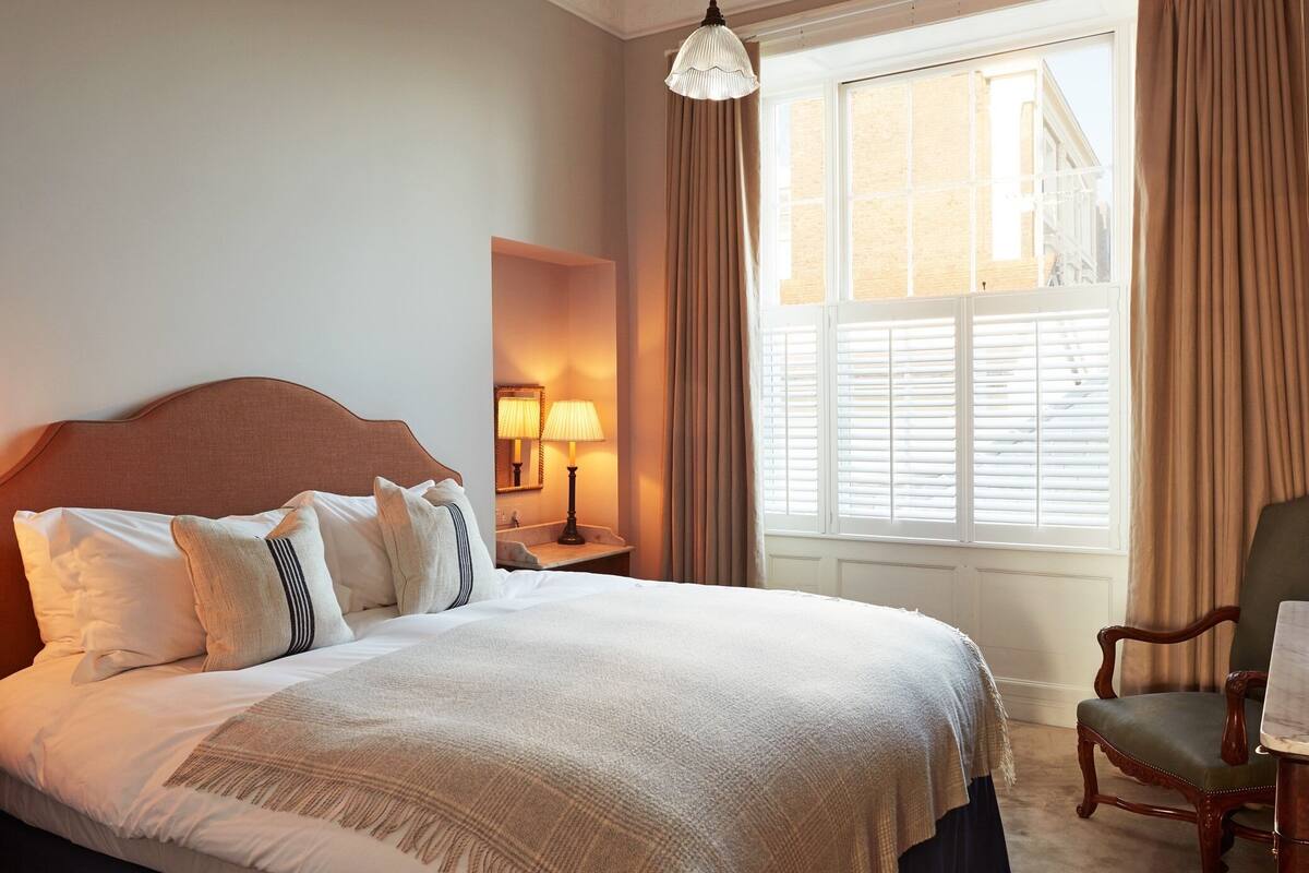 A neatly made bed with white linens, a wooden headboard, and a bedside lamp. A chair is positioned near a window with curtains.