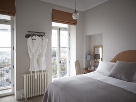 A hotel room with a large bed, a radiator, a chair, a desk with a lamp, and a window with a view of the city.